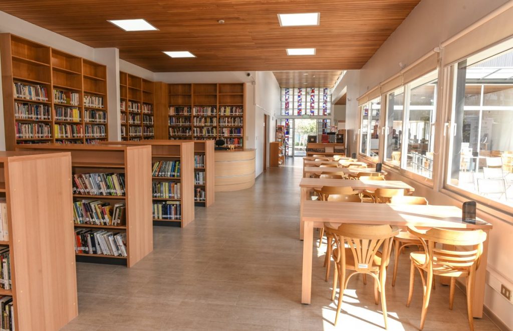 Cafetería de colores beage y café claro, con ventanas a la derecha, mesas y sillas y a la izquierda libreros altos y medianos. 