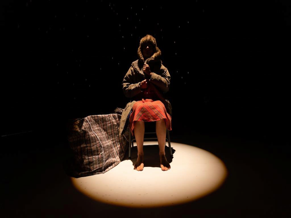 Actriz sentada en un escenario oscuro, iluminada por un foco arriba suyo. 