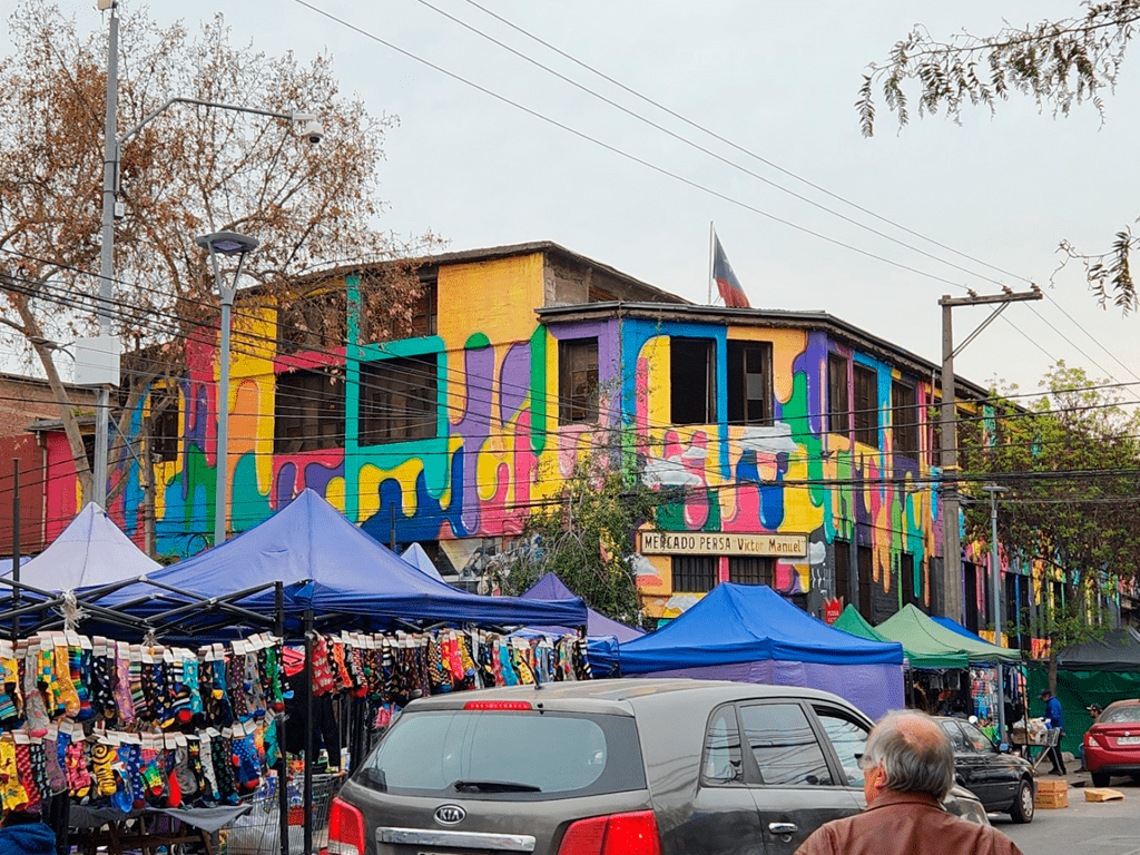 mercado callejero con puestos de venta y carpas coloridas. Los puestos exhiben una variedad de artículos. Al fondo, un edificio con un mural multicolor que presenta formas abstractas en tonos brillantes de amarillo, azul, verde, rosa y morado. El edificio con un letrero que dice Mercado Persa Víctor Manuel