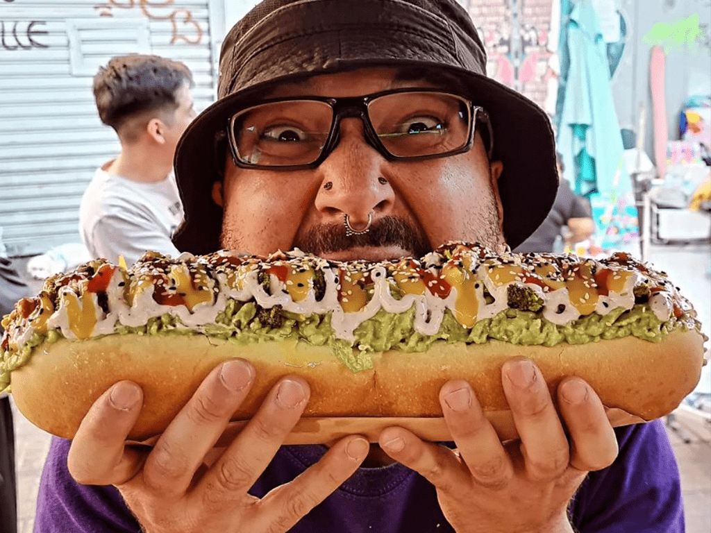 Persona sosteniendo un Completo vegano grande con palta, tomates, sésamo y salsas.