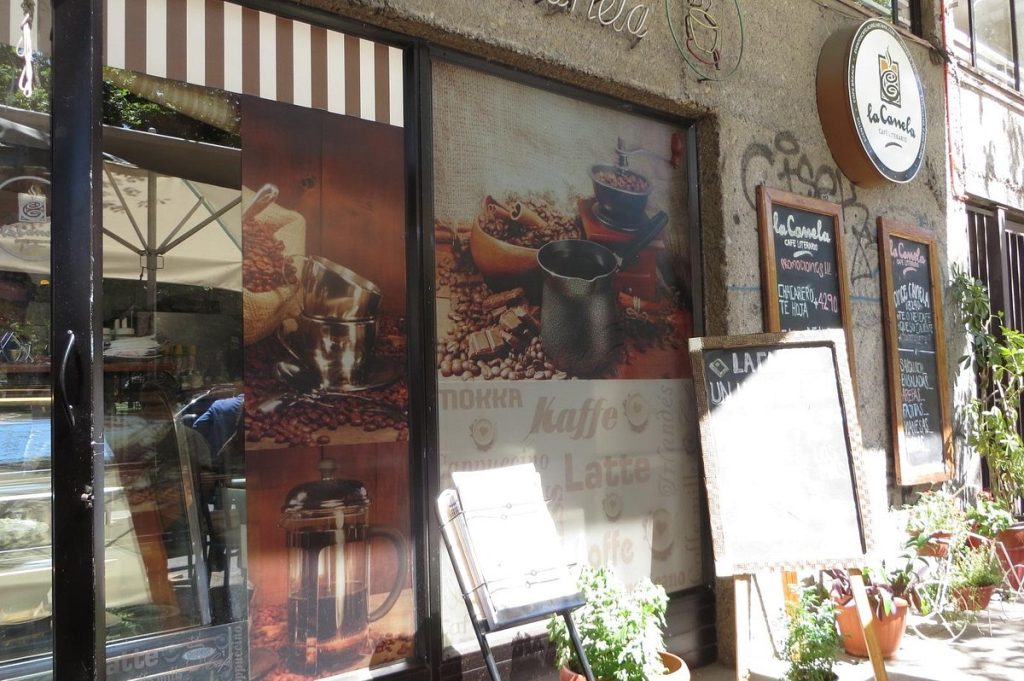 Imagen desde afuera de cafetería "La Canela", donde se puede apreciar un cartel con su menú y un papel mural de granos de café y cafeteras. 