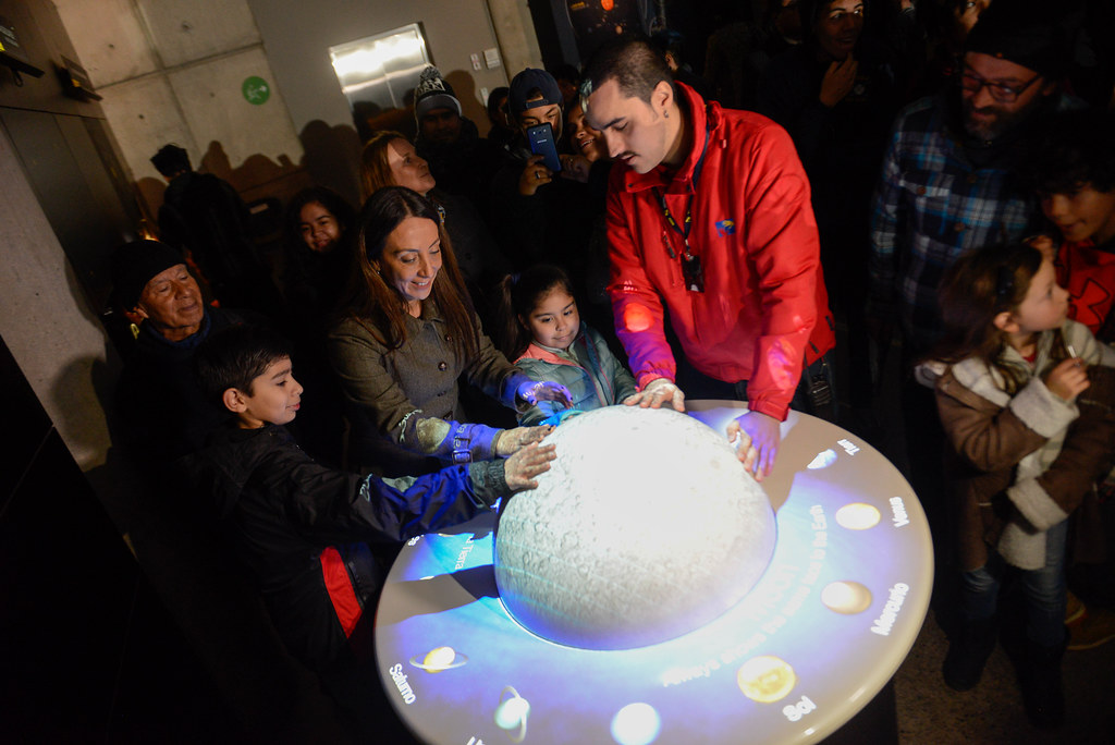 Adultos y niños ponen sus manos sobre un sistema solar en el Museo Interactivo Mirador. Licencia Creative Commons.