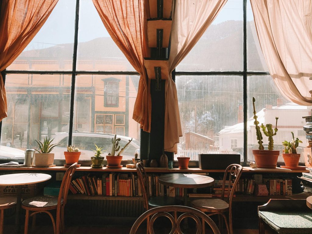 La fotografía muestra dos mesas en una cafetería, con sillas, al lado de un ventanal y bajo este un librero lleno de libros.