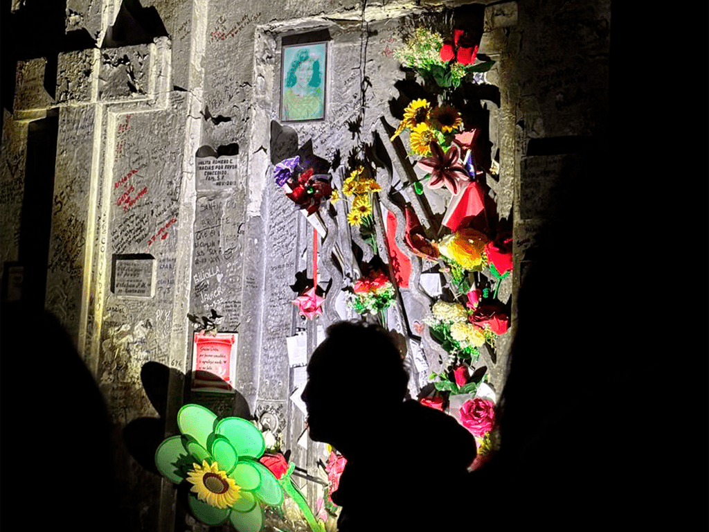 Tumba de Orlita decorada con flores frescas y cintas coloridas, situada en un cementerio. Las paredes de piedra alrededor están llenas de inscripciones y mensajes personales, frente a esto la sombra del guía del tour paranormal.