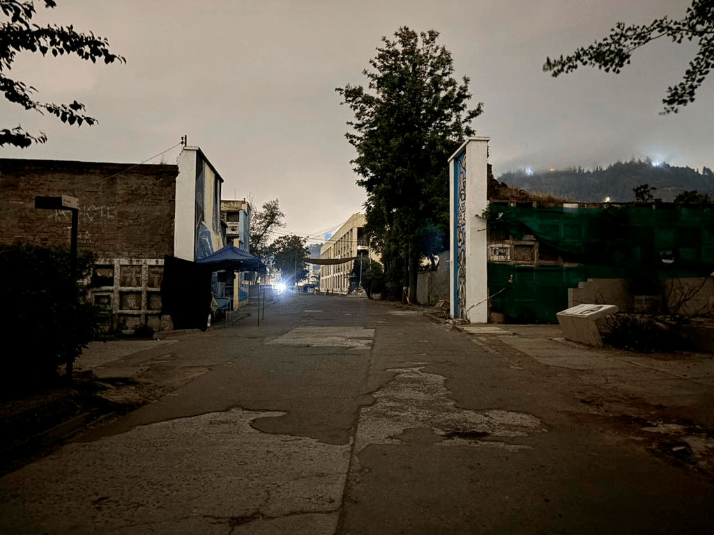 Pasillo en el Cementerio General, con iluminación tenue y rodeado de nichos y placas conmemorativas. A la izquierda, se observa una hilera de nichos decorados con flores y otros objetos. A la derecha, otro muro con más nichos y placas con inscripciones.