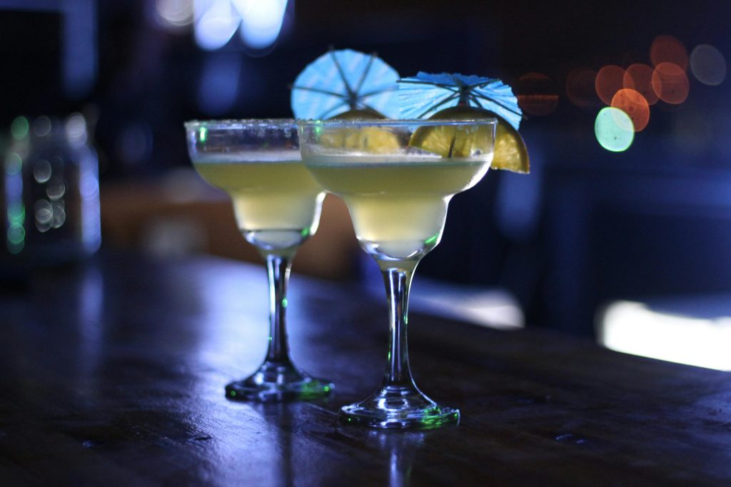 Dos cócteles margarita servidos en copas de vidrio con bordes escarchados, decorados con rodajas de limón y sombrillas azules en un ambiente nocturno con luces difusas
