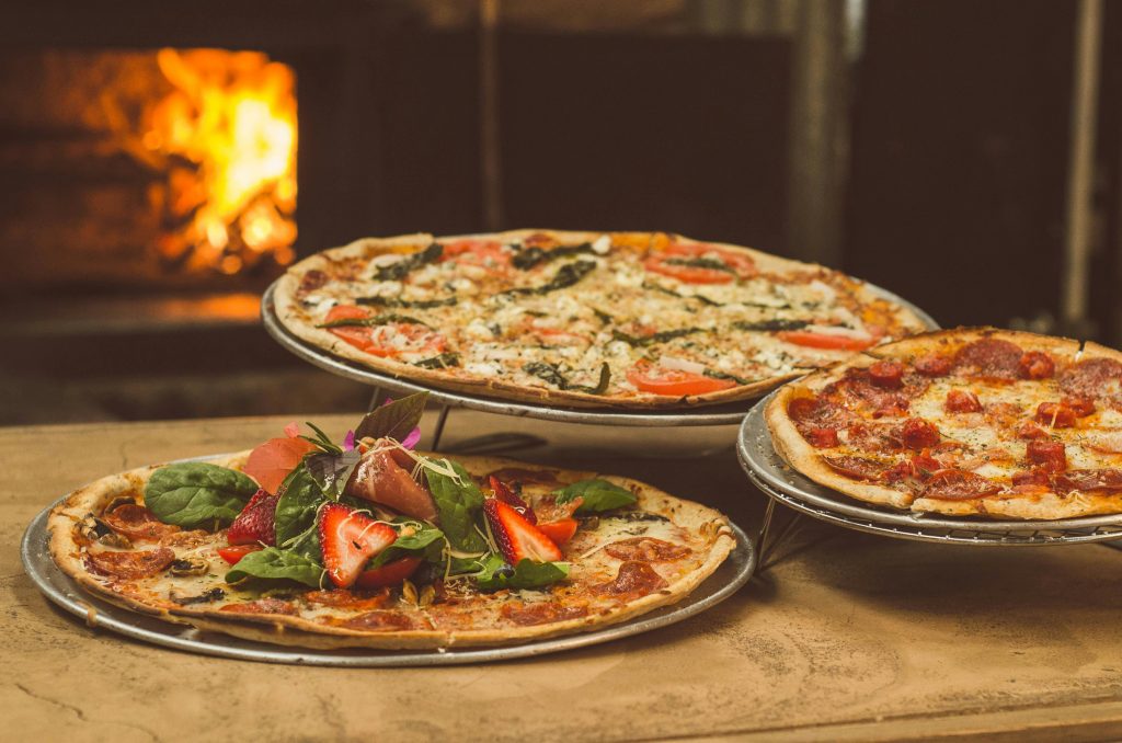 Tres pizzas artesanales servidas sobre bandejas metálicas: una con pepperoni, otra con vegetales frescos como espinaca y tomate, y una decorada con fresas y hojas verdes, colocadas sobre una mesa de madera frente a un horno de leña encendido que aporta un ambiente cálido.