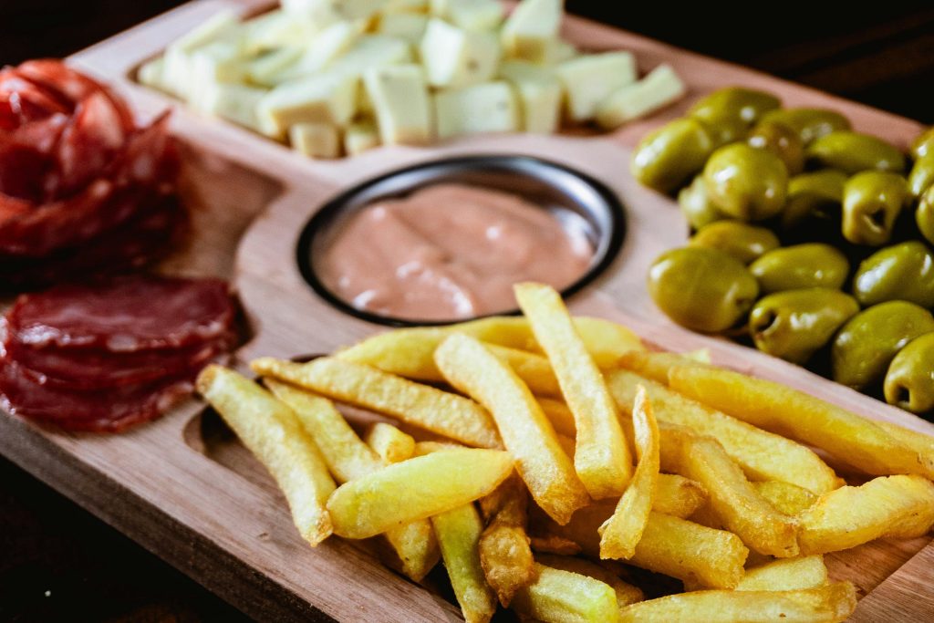 Tabla de aperitivos con papas fritas, aceitunas verdes, trozos de queso, salame y salsa rosada en un recipiente central, perfecta para compartir en reuniones o eventos sociales. Ideal para amantes de los snacks y comidas rápidas