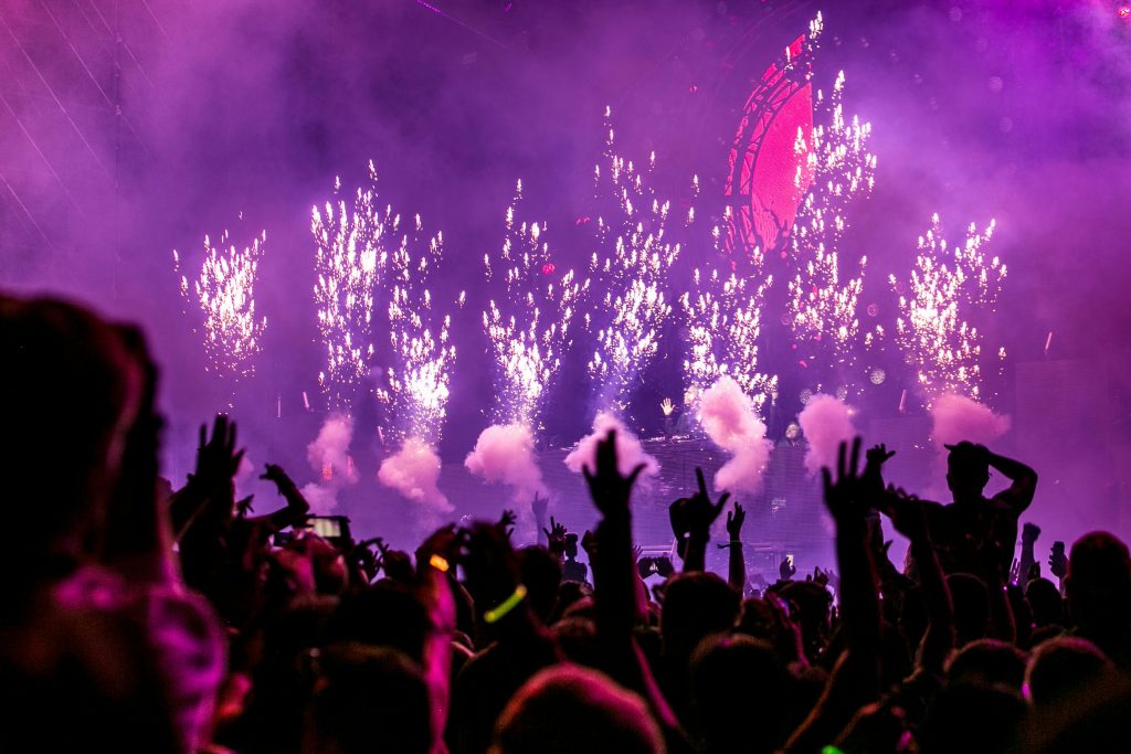 La imagen muestra una escena de un concierto nocturno con un espectáculo de fuegos artificiales y humo en el escenario. Las luces predominantes son de color púrpura, creando una atmósfera vibrante y festiva. El público está de pie, con las manos levantadas, disfrutando de la música y el ambiente. En el fondo se observa una estructura circular iluminada, donde parece estar el DJ o artista actuando. La energía del evento es palpable, con el juego de luces, fuegos artificiales y la multitud participando activamente.