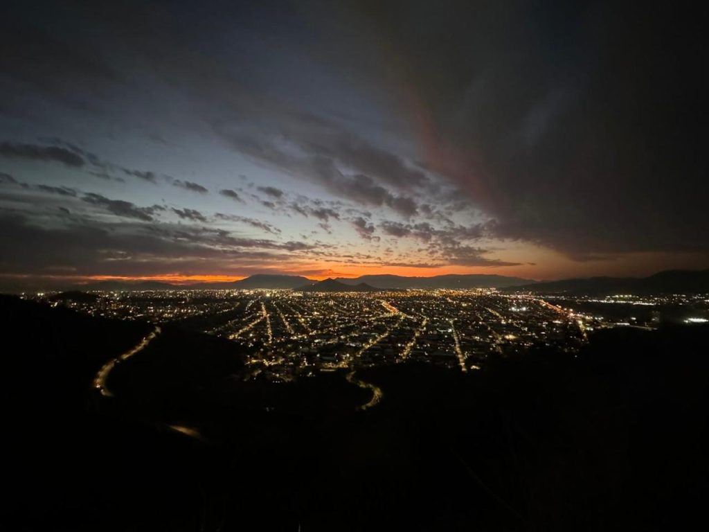Fotografía nocturna de autoría propia en la que aparece una vista panorámica de Santiago iluminada, con un atardecer ya extinguiéndose desde el punto de vista de la cumbre del cerro El Carbón.