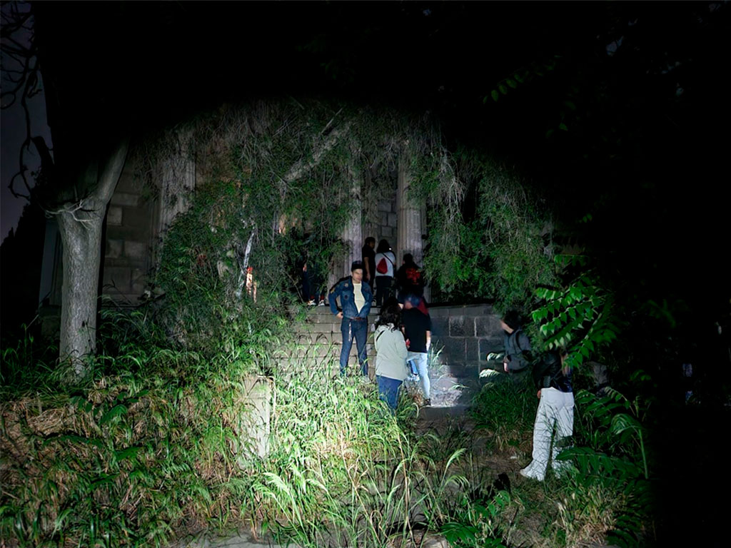 Visitantes explorando una antigua estructura cubierta de vegetación en un entorno nocturno, con linternas iluminando el camino.
